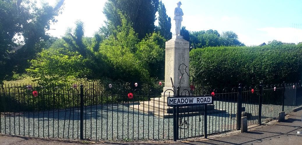 image of the Catshill war memorial site, including pillar with names of the fallen, statue of a soldier, meadow road sign and black metal fence decorated with poppies 